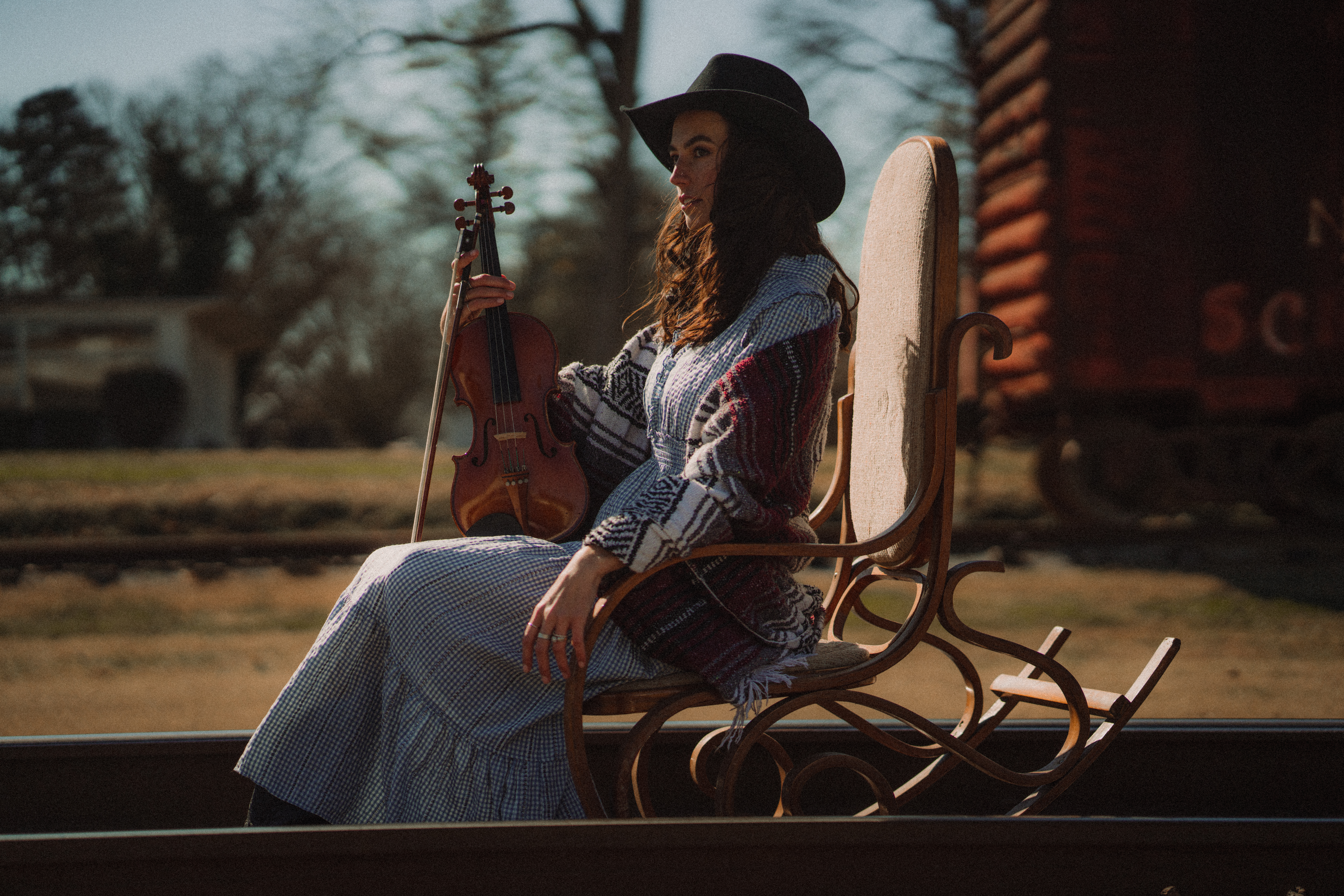 Woman in cowboy hat and blue dress sits in rocking chair on train tracks with a violin in her hand.