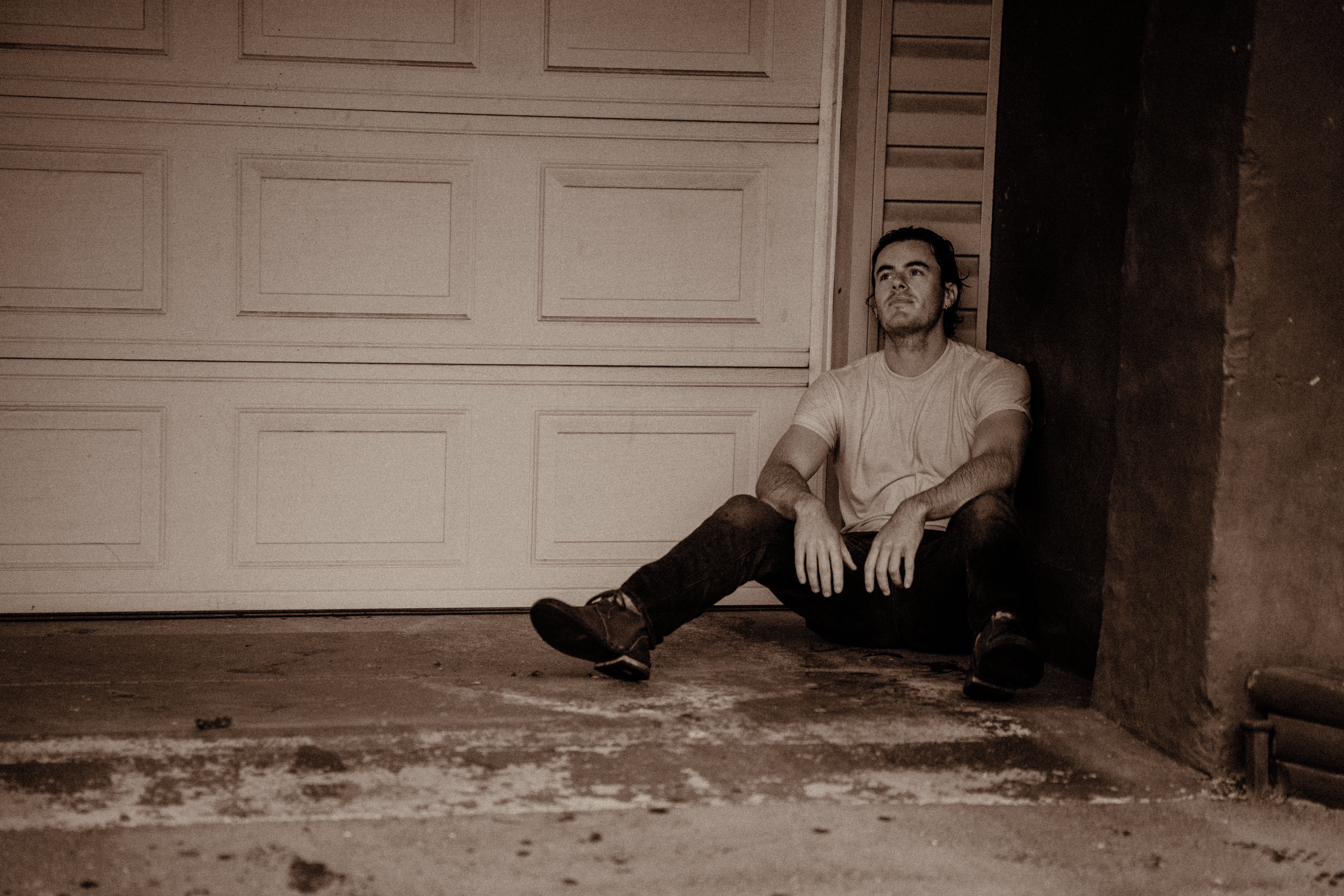 Guitar player soaked with rain sits in a corner by a garage door with his arms hanging on his knees.